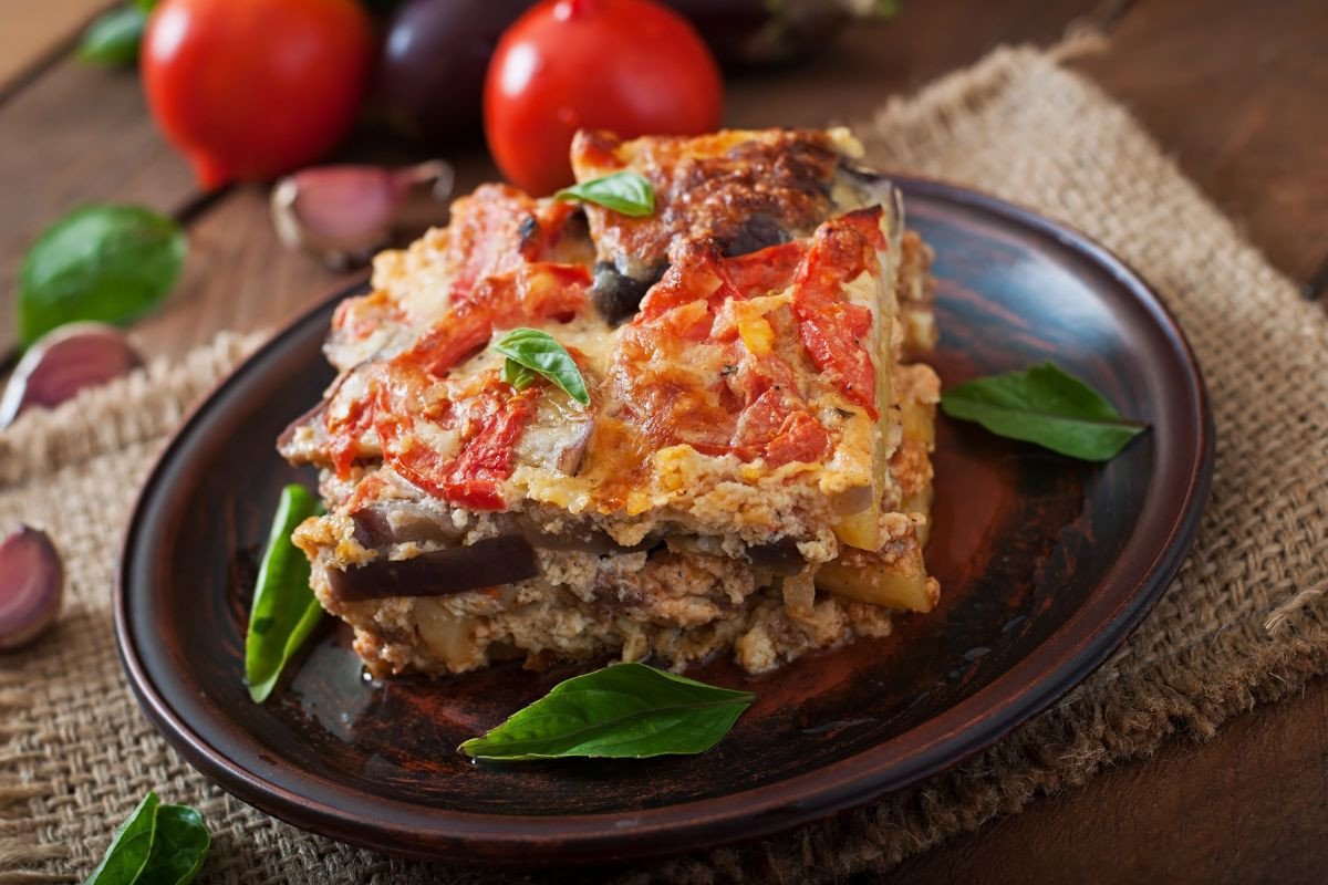 A slice of moussaka on a dark plate, garnished with fresh basil leaves, with tomatoes and garlic in the background.